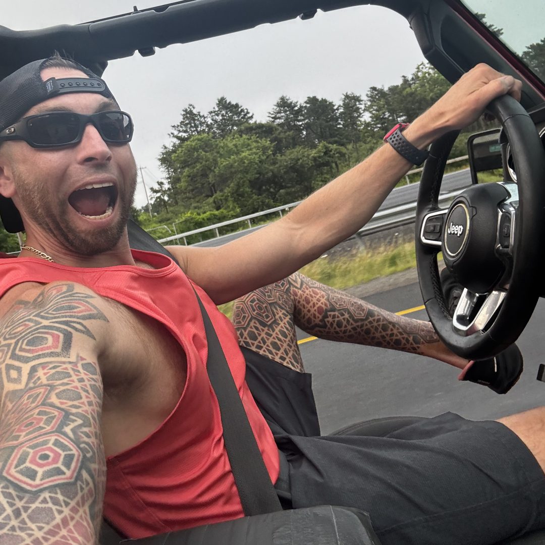 Adam driving his Jeep with the top off, mid-shout and grinning, tattooed arm on the wheel.