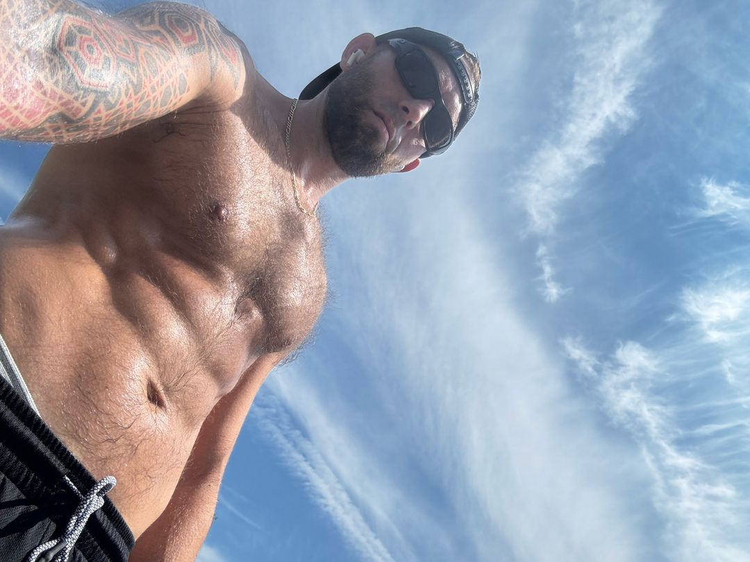 Low-angle portrait of Adam outdoors against a blue sky, shirtless after a workout, tattooed sleeve visible.
