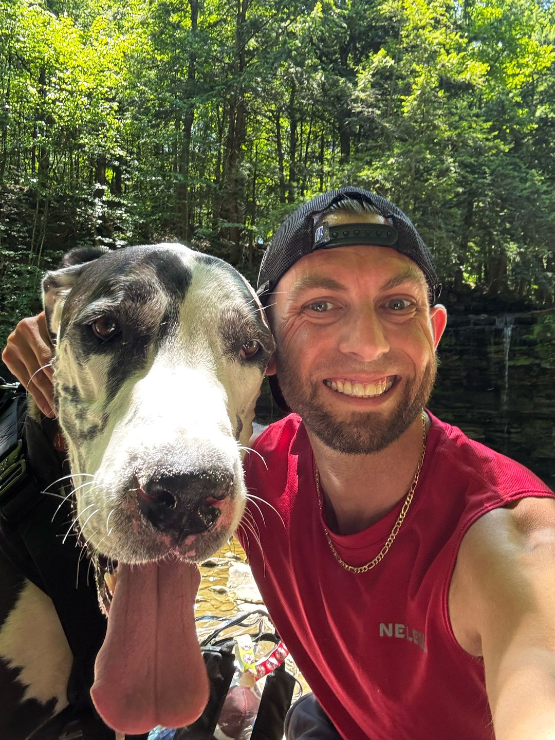 Adam, grinning at the camera in a backwards cap, beside his Great Dane Smoke at a creek in the woods.