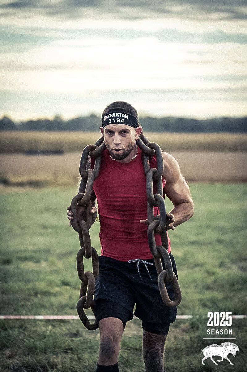 Adam mid-race at a Spartan event, carrying a heavy iron chain across his shoulders through a field.