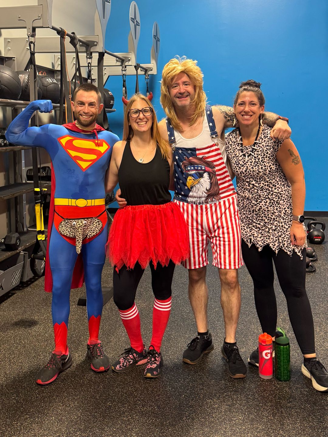 Adam in a full Superman costume, flexing, with his arm around three friends in costume at his gym.
