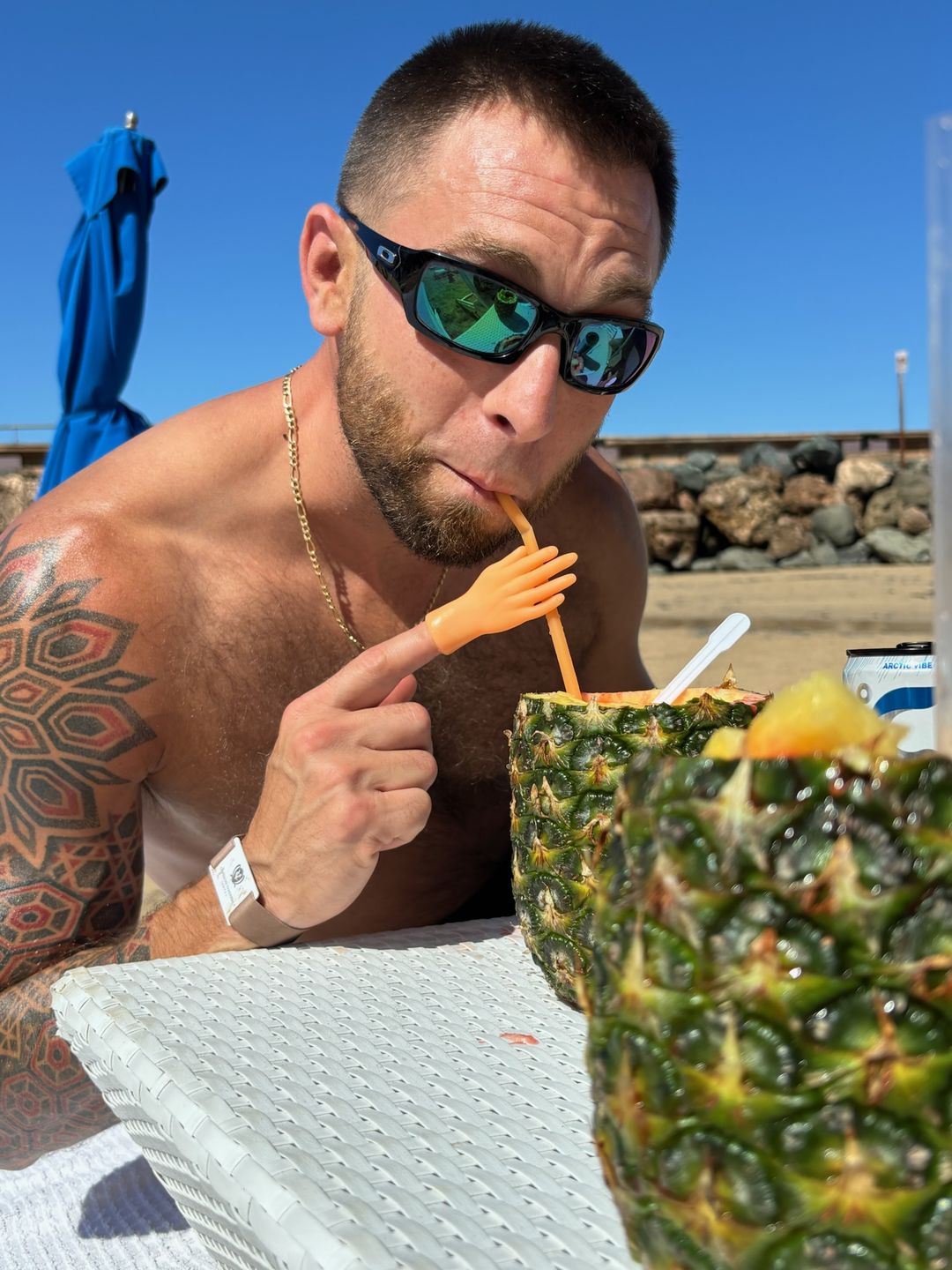 Adam on a beach in sunglasses, drinking from a hollowed pineapple through a straw held by a tiny plastic doll hand on his finger.