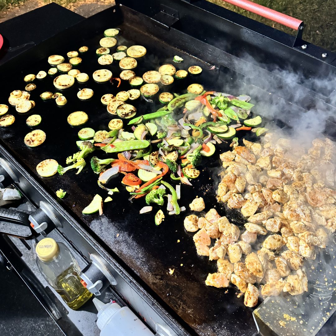 Chicken, zucchini, peppers, broccoli and onions cooking on an outdoor griddle.